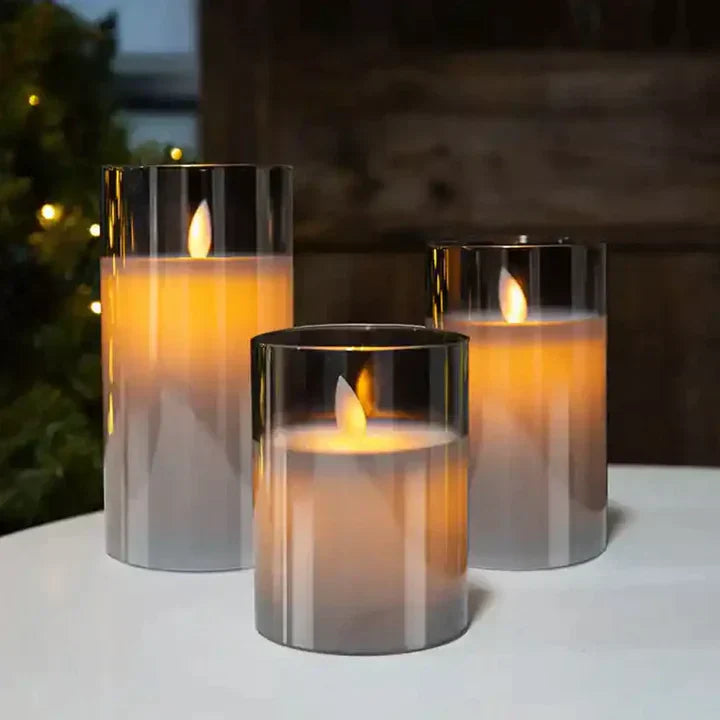 Drei LED-Kerzen in Glaszylindern, warmweiß leuchtend, auf weißem Tisch, dekorative Beleuchtung.