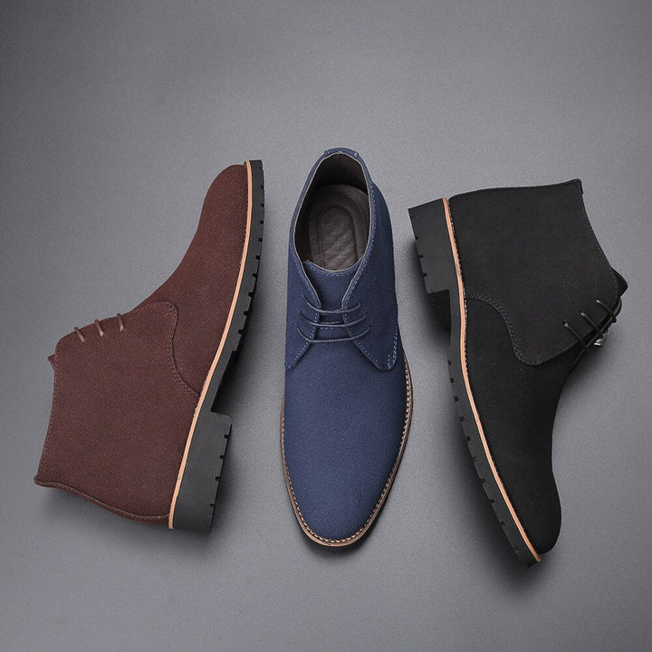 Drei Herren-Chelsea-Stiefel aus Wildleder in Braun, Blau und Schwarz auf grauem Hintergrund.