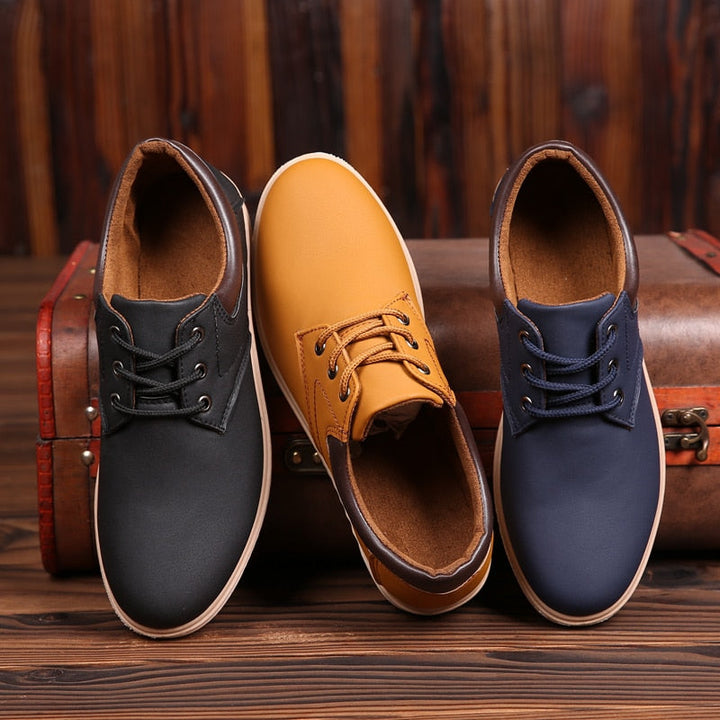 Drei Paar Herren-Ledersneaker in Schwarz, Gelb und Blau auf Holzhintergrund vor Koffer.