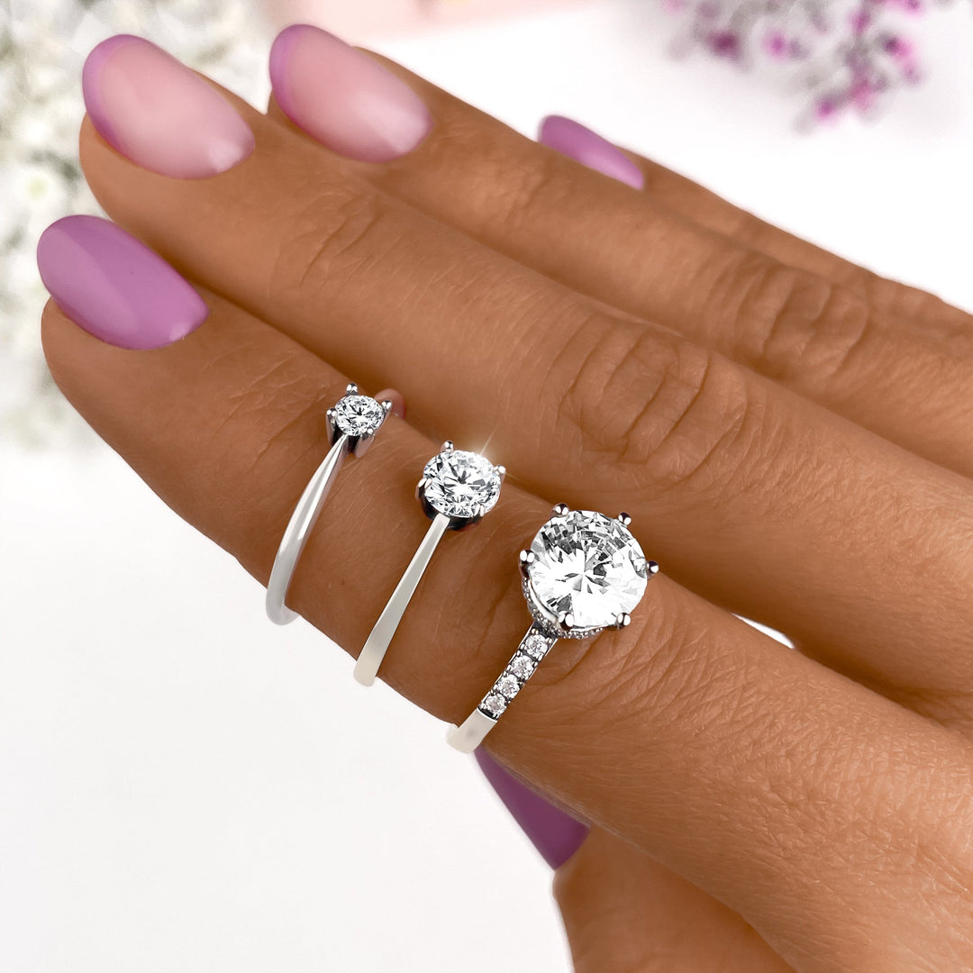 Drei silberne Verlobungsringe mit Diamanten auf Hand, lila Nagellack, elegantes Schmuckdesign.