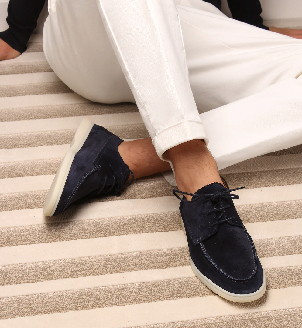 Dunkelblaue Wildleder-Sneaker für Herren, getragen mit weißen Hosen, auf gestreiftem Teppich.