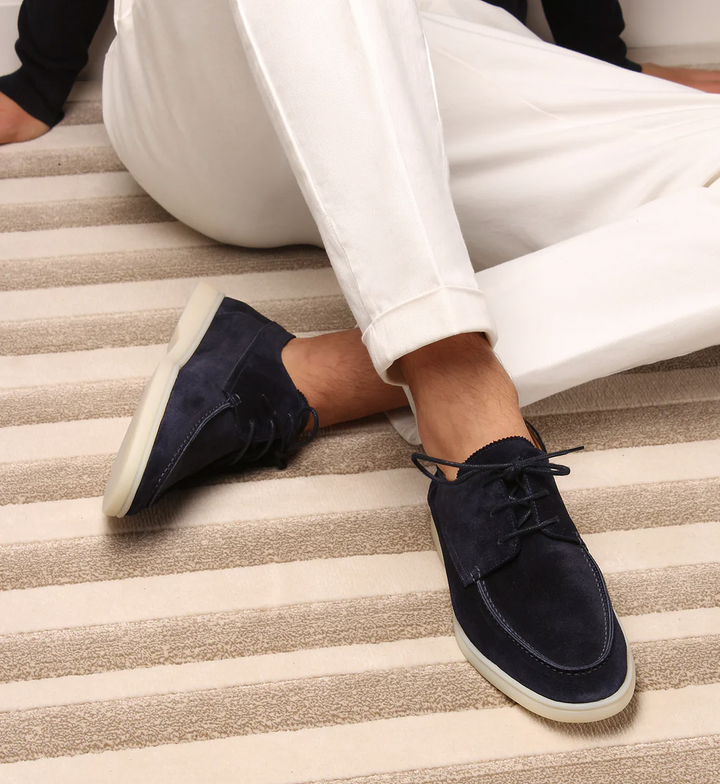 Dunkelblaue Wildleder-Sneaker für Herren, getragen mit weißen Hosen, auf gestreiftem Teppich.