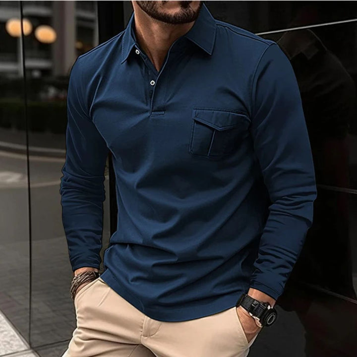Mann in dunkelblauem Langarm-Poloshirt, Brusttasche, beiger Hose, Armbanduhr, urbaner Hintergrund.