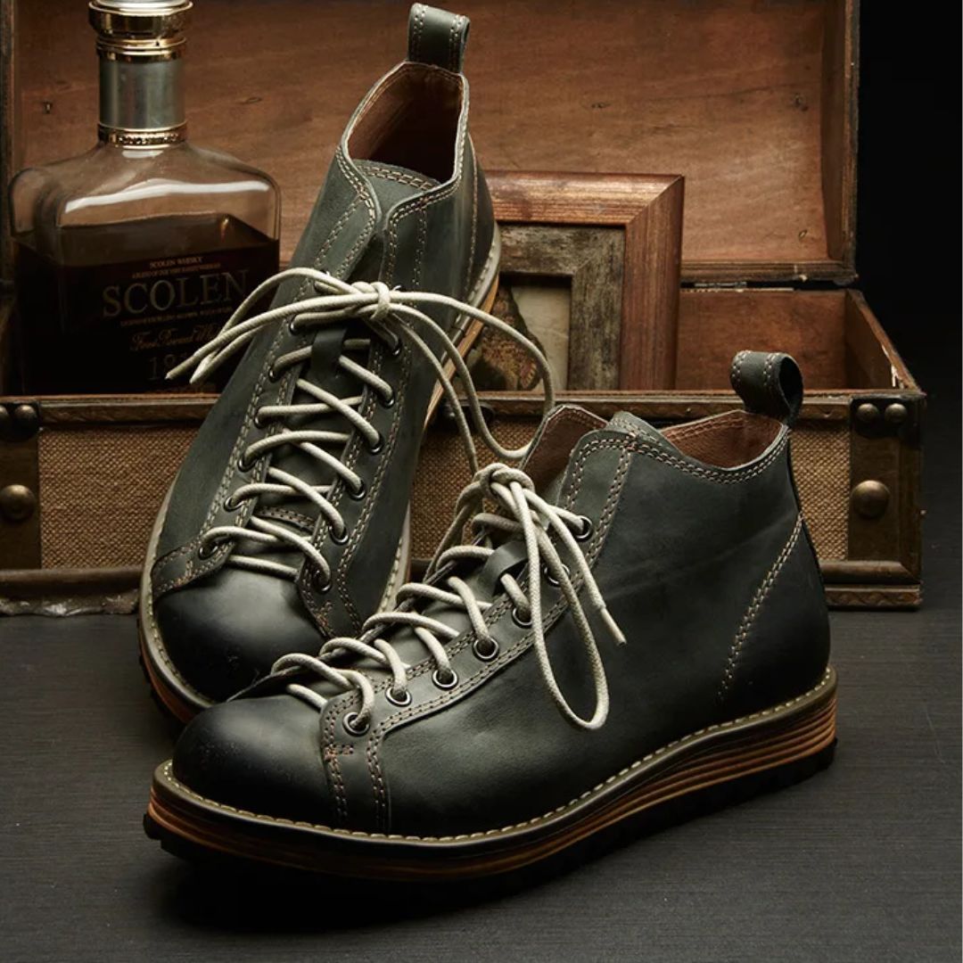 Graue Lederstiefel mit Schnürsenkeln, Vintage-Stil, vor Holzkiste und Flasche.