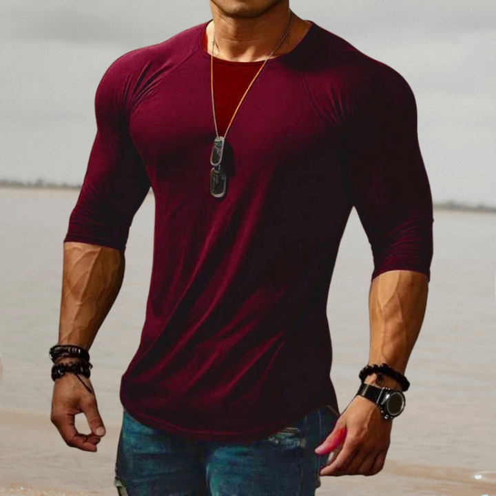 Mann in bordeauxrotem Langarmshirt, muskulös, mit Halskette und Armbändern am Strand.