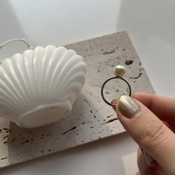 Hand hält silbernen Ring mit Perle, Muschel-Dekoration im Hintergrund, eleganter Schmuck.