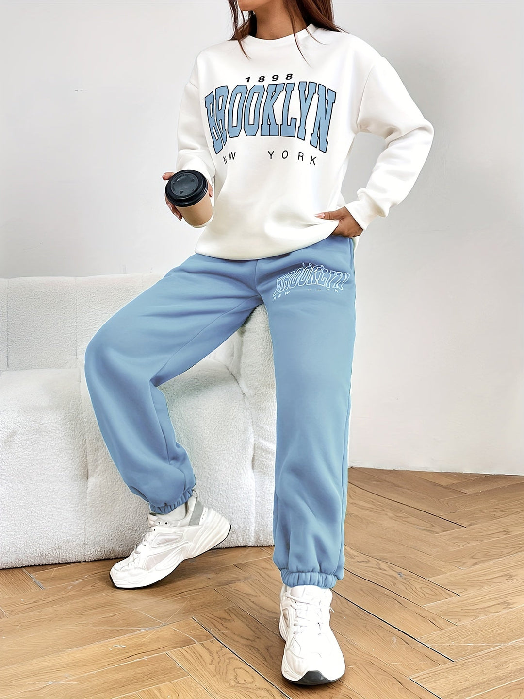 Frau in weißem "Brooklyn" Sweatshirt und blauer Jogginghose, hält schwarzen Kaffeebecher.