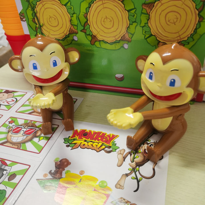 Zwei braune Affen-Spielzeugfiguren vor einem bunten Brettspiel, "Monkey Toss" Verpackung.