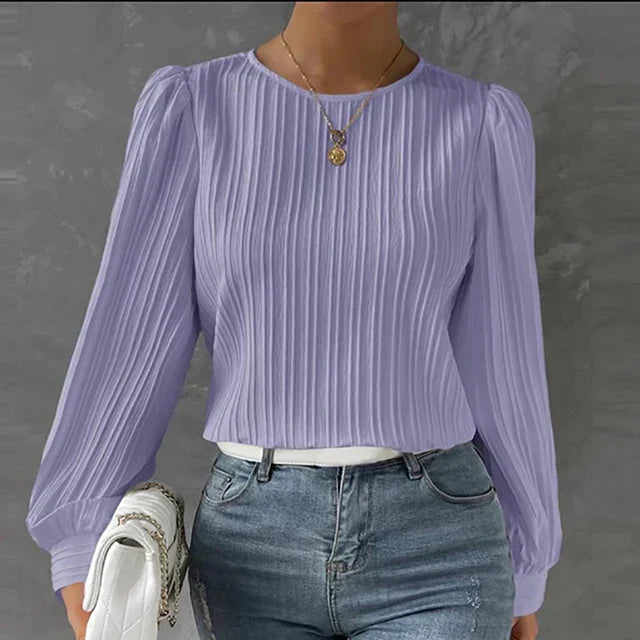 Frau in lila plissierter Bluse mit langen Ärmeln, kombiniert mit blauer Jeans und weißer Tasche.