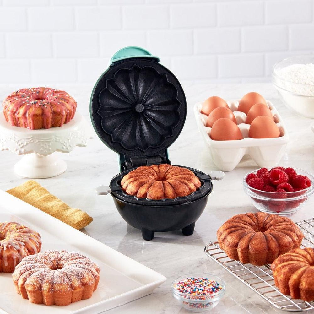 Elektrische Kuchenbackmaschine, schwarz, mit gebackenem Kuchen, umgeben von Eiern und Zutaten.