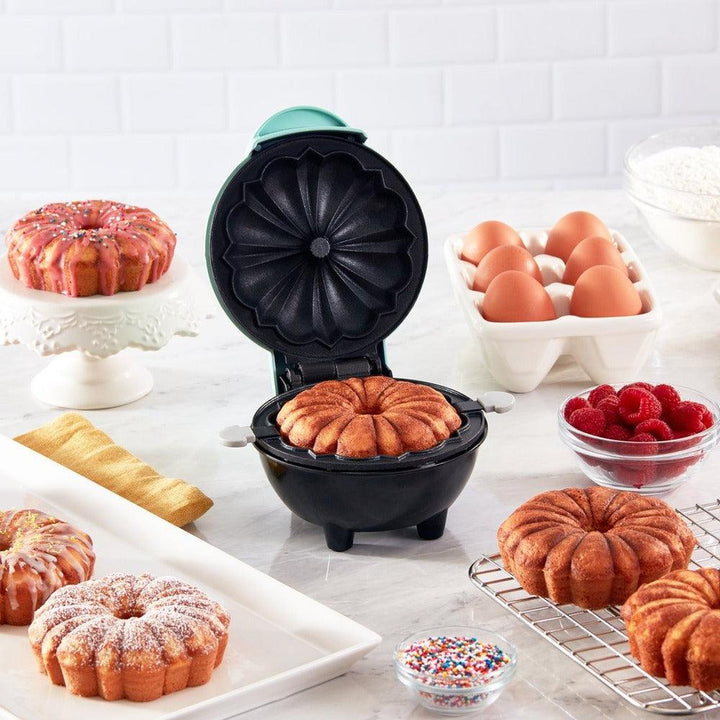 Elektrische Kuchenbackmaschine, schwarz, mit gebackenem Kuchen, umgeben von Eiern und Zutaten.