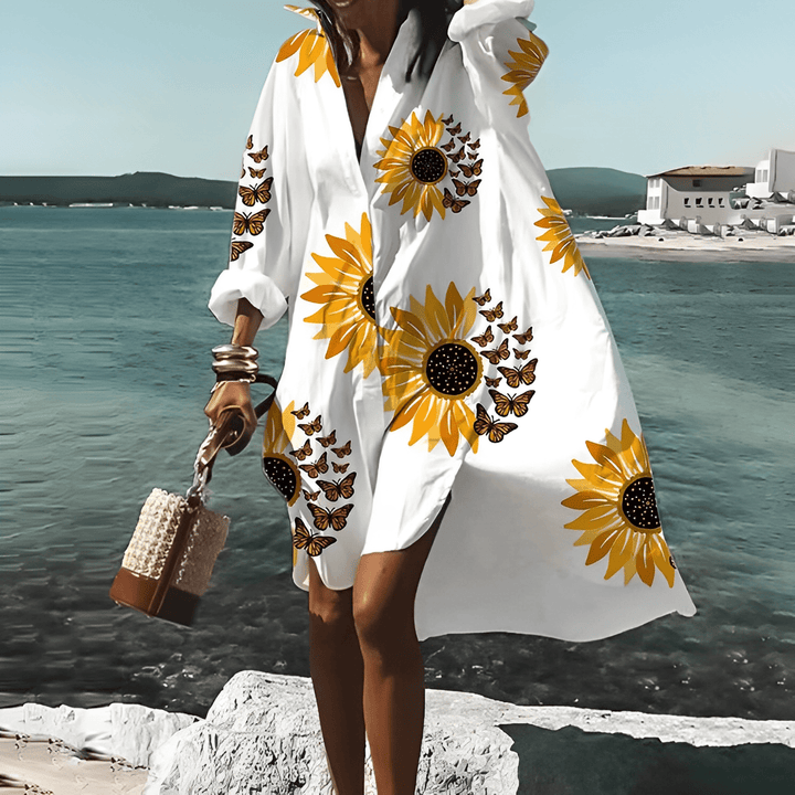 Frau im weißen knielangen Kleid mit Sonnenblumenmuster am Strand.