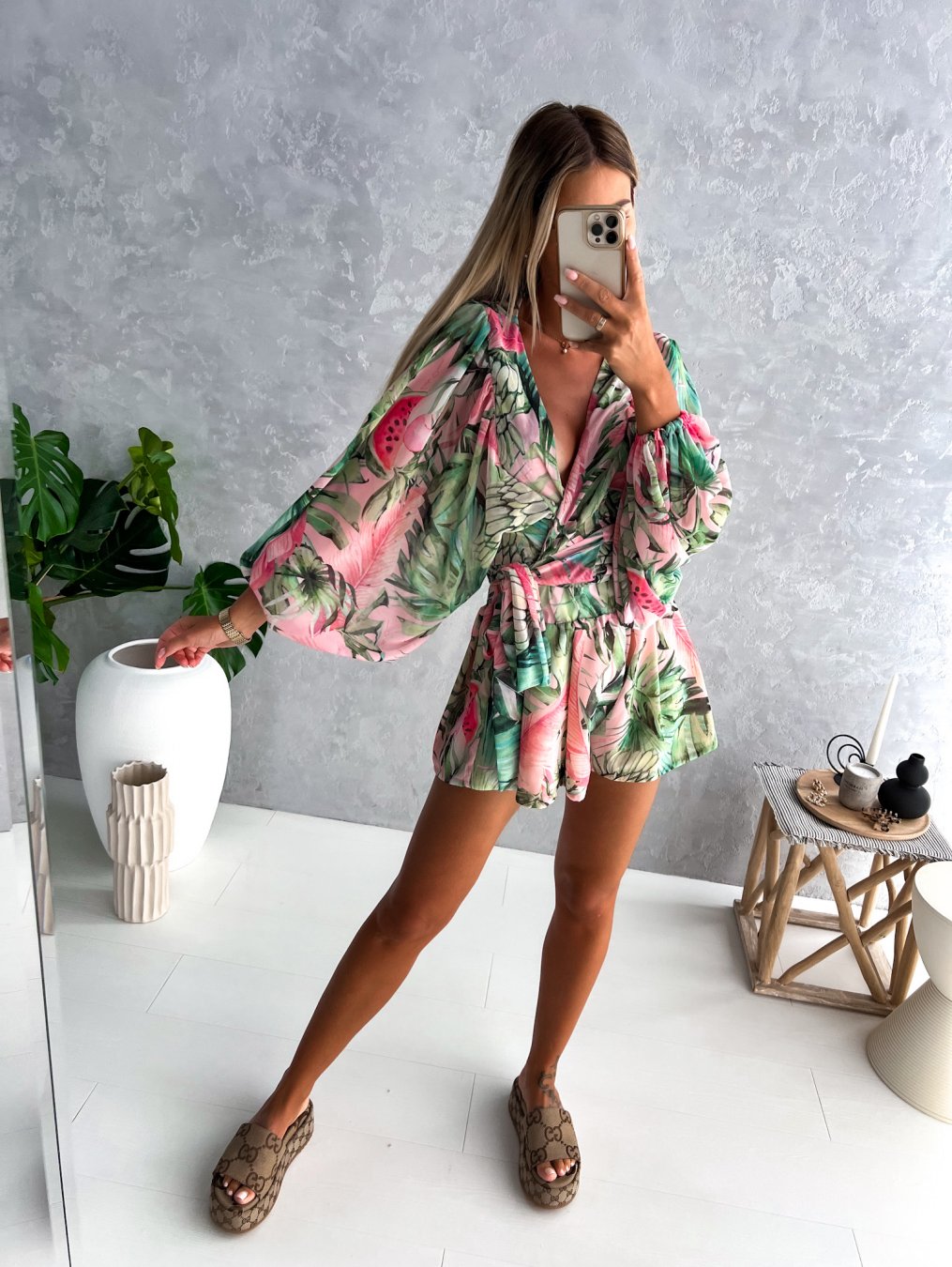 Frau im grünen-pinken Sommerkleid mit tropischem Muster, steht in modernem Wohnzimmer.