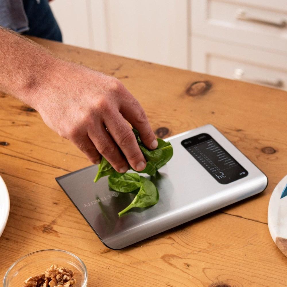 Digitale Küchenwaage aus Edelstahl mit Hand, die Spinat wiegt, auf Holztisch.