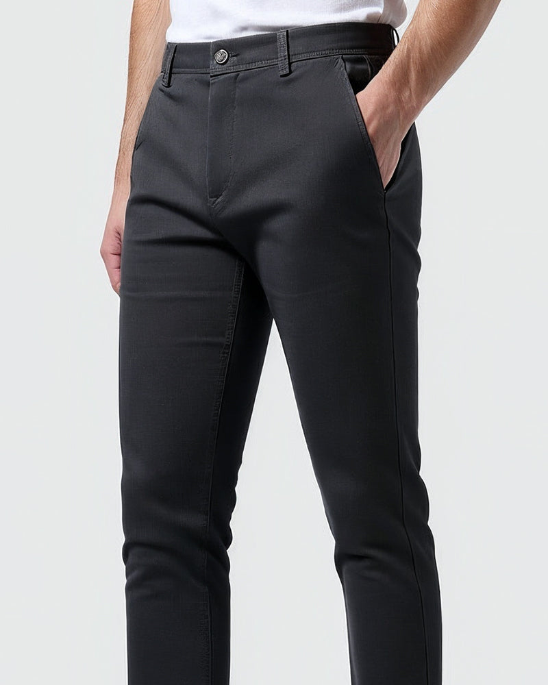 Detail pasu a přední kapsy tmavých chino kalhot, knoflík a poklopec zblízka, model má jednu ruku v kapse. 10