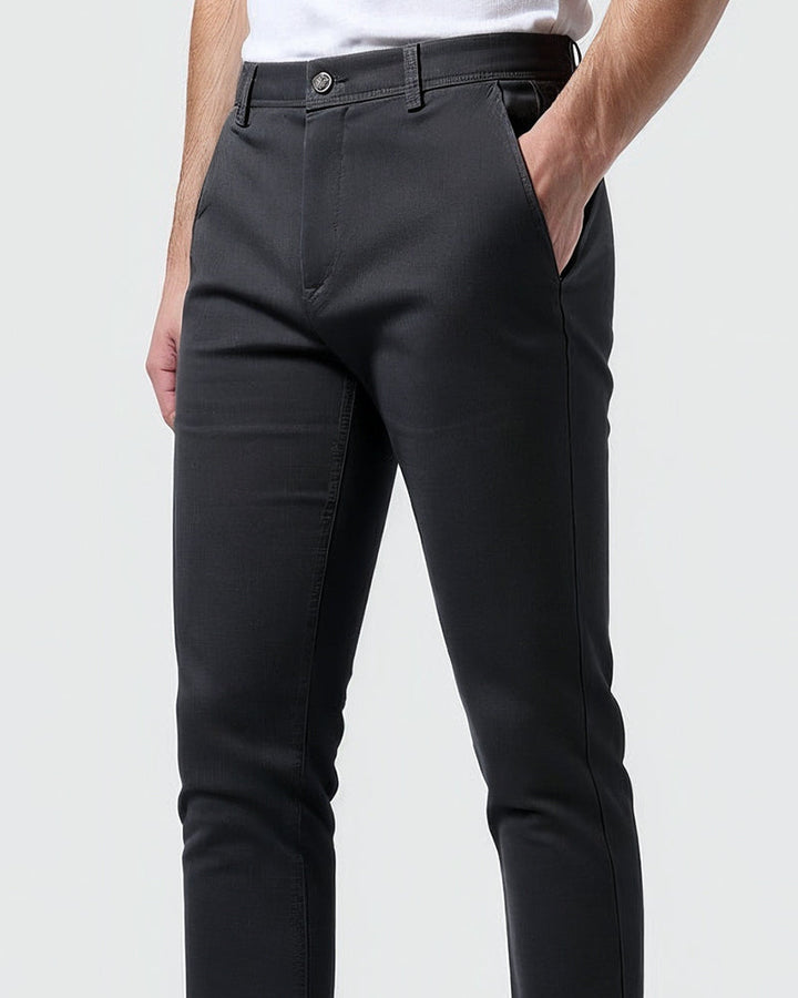 Detail pasu a přední kapsy tmavých chino kalhot, knoflík a poklopec zblízka, model má jednu ruku v kapse. 10