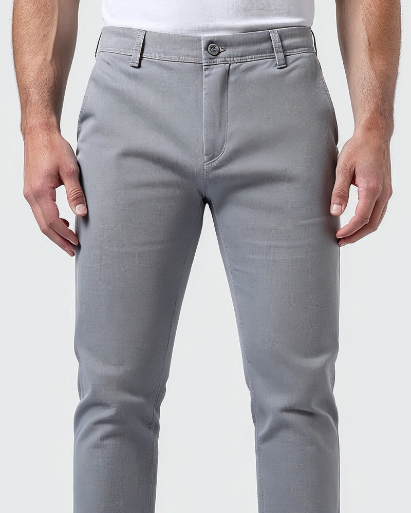 Detail pasu světle šedých chino kalhot zepředu: knoflík, poutka na opasek, poklopec a švy, model má ruce volně podél těla. 5