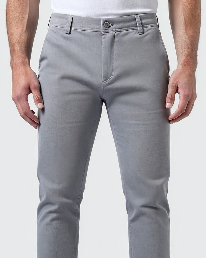 Detail pasu světle šedých chino kalhot zepředu: knoflík, poutka na opasek, poklopec a švy, model má ruce volně podél těla. 5
