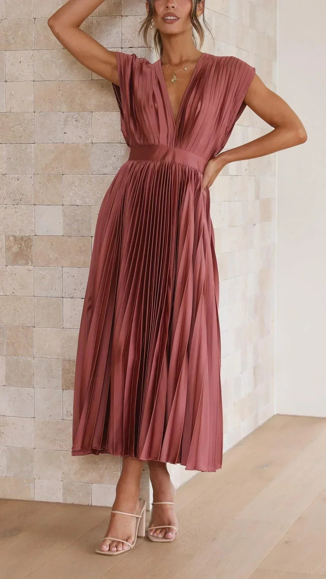 Frau in rotem, plissiertem Maxikleid aus Satin, ärmellos, elegantes Design, vor Steinwand.