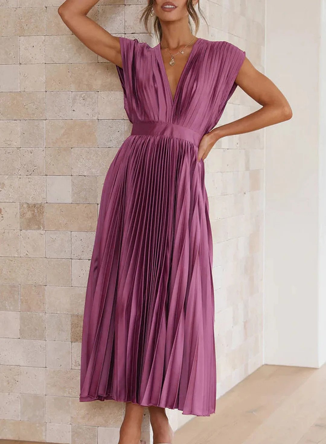 Frau in lila plissiertem Kleid, ärmellos, V-Ausschnitt, elegantes Design, vor beiger Wand.