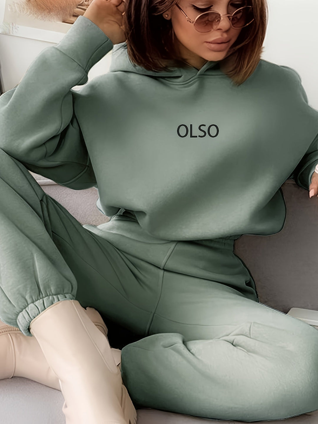 Frau in grünem OLSO-Hoodie und Jogginghose, trägt Sonnenbrille, sitzt auf Sofa.