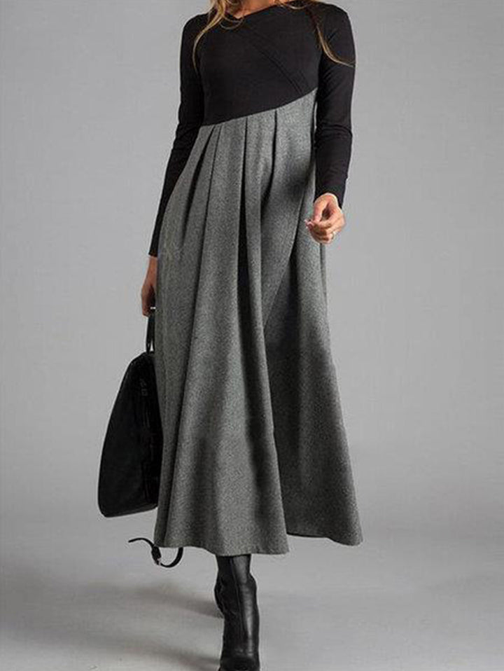 Frau in langem, grauem Maxikleid mit schwarzen Ärmeln und Stiefeln, hält schwarze Tasche.