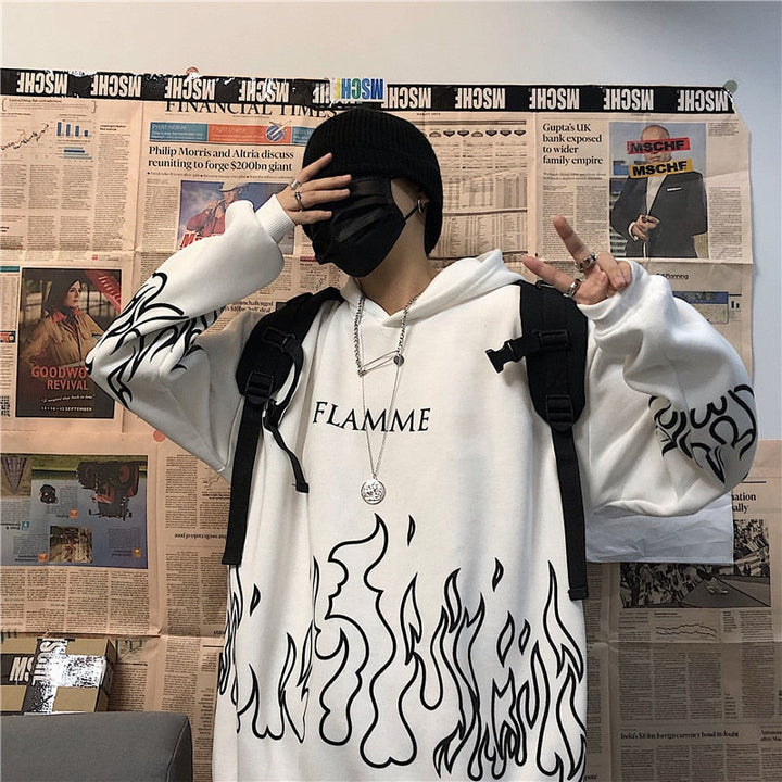 Person in weißem Hoodie mit Flammenmuster, schwarzer Mütze und Maske, vor Zeitungswand.