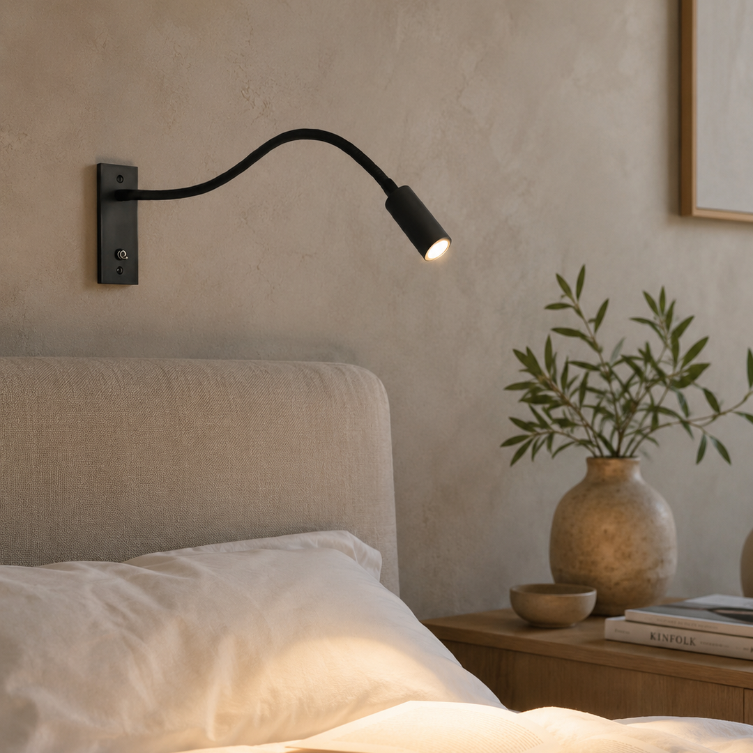 Černá čtecí lampa na štukové zdi nad postelí, svítí teplým světlem na rozečtenou knihu a polštáře. 3