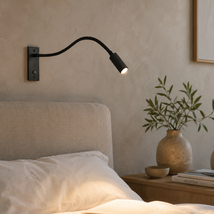 Černá čtecí lampa na štukové zdi nad postelí, svítí teplým světlem na rozečtenou knihu a polštáře. 3