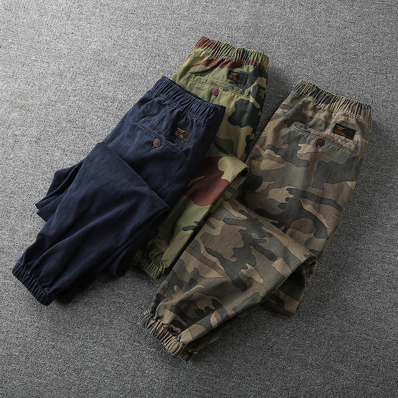 Drei Herren-Cargohosen, Camouflage und Blau, elastischer Bund, auf grauem Hintergrund.