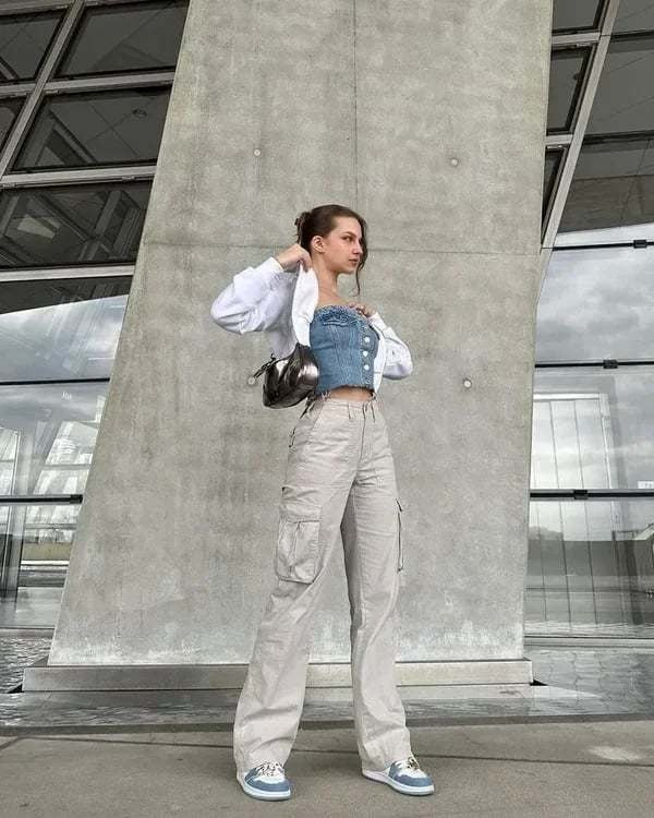 Frau in weißem Hemd, blauer Jeansweste, beigen Cargohosen, blauen Sneakers vor moderner Architektur.