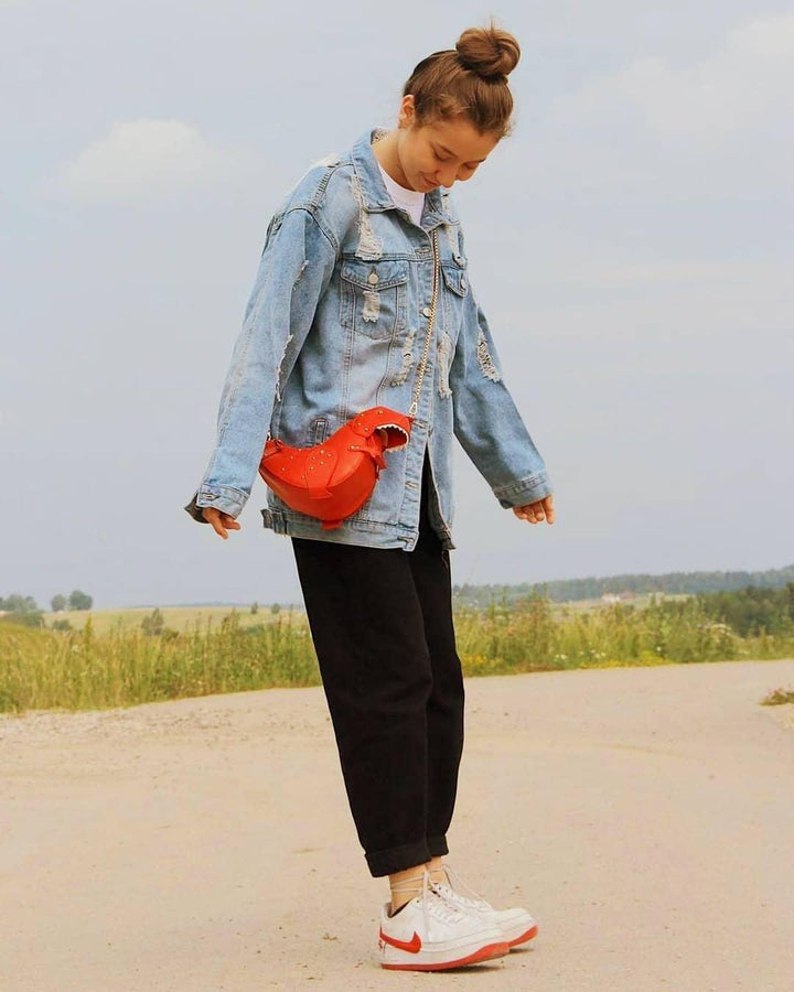Frau in blauer Jeansjacke, schwarzer Hose, roter Tasche und weißen Sneakers im Freien.
