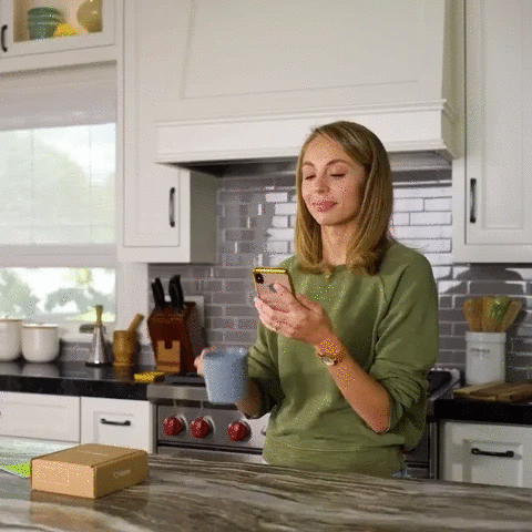 Frau in Küche mit Smartphone und Kaffeetasse, moderne Einrichtung, graue Fliesen, Holzarbeitsplatte.