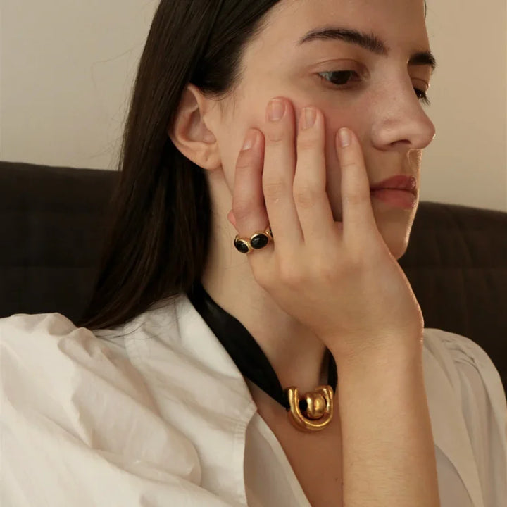 Frau mit schwarzem Ring und goldener Halskette, weißes Hemd, stilvolles Schmuckdesign.