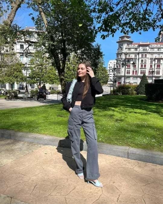 Frau in schwarzer Jacke und grauer Hose im Park, sonniger Tag, moderne Stadtumgebung.