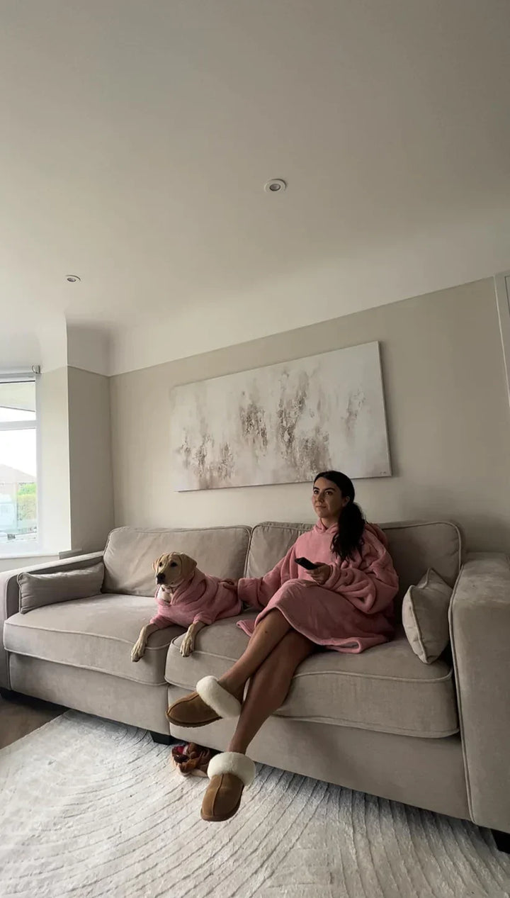 Frau und Hund in rosa Decken auf grauem Sofa, gemütliches Wohnzimmer, entspannte Atmosphäre.