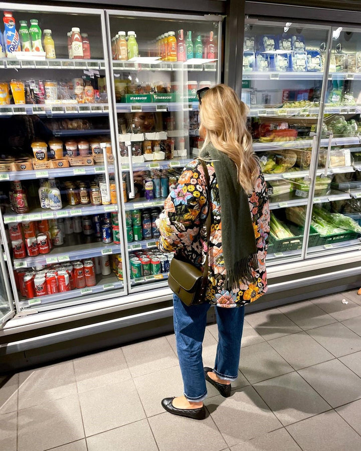 Frau im Supermarkt vor Kühlregal, trägt bunte Jacke, Jeans, schwarze Schuhe, grüne Tasche.