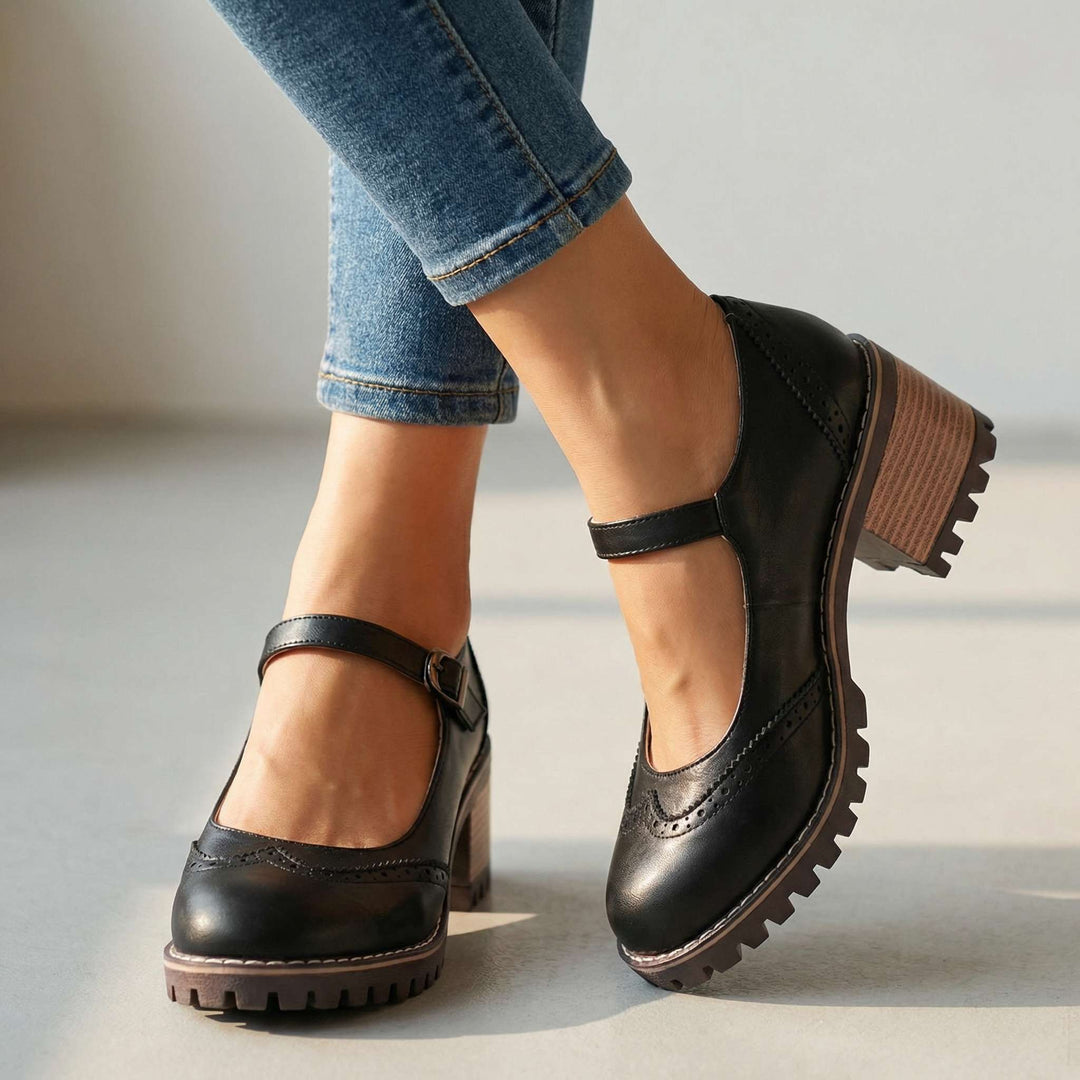 Černé boty s perforovanou špičkou a páskem přes nárt, nasazené na nohou v džínách, detail v interiéru se světlým pozadím. 1