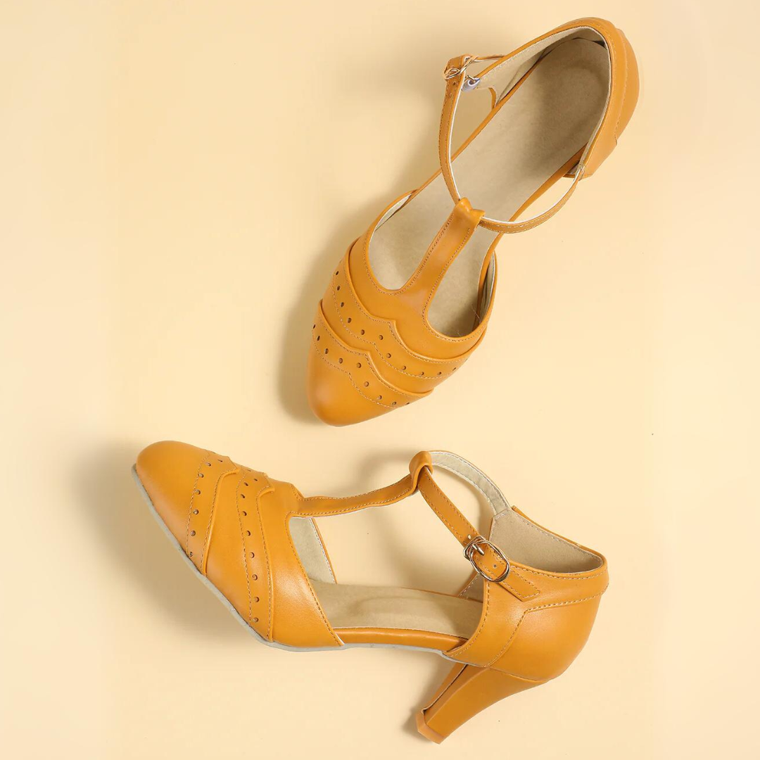 Gelbe Damen-Tanzschuhe mit Absatz, Riemchen und Lochmuster auf beigem Hintergrund.