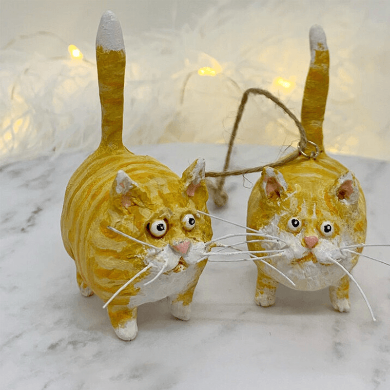 Zwei gelbe Katzen-Weihnachtsornamente aus Keramik mit Schnur auf Marmortisch.