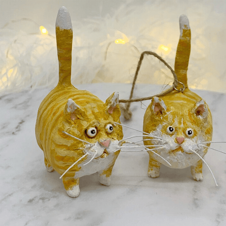 Zwei gelbe Katzen-Weihnachtsornamente aus Keramik mit Schnur auf Marmortisch.