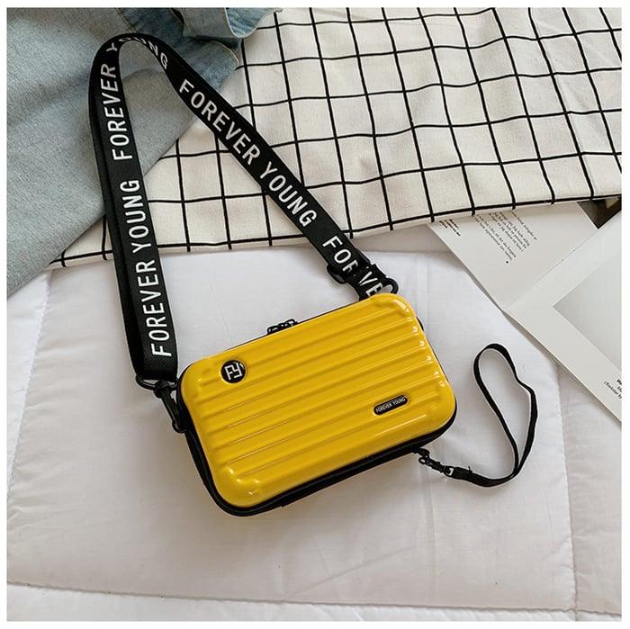 Gelbe Mini-Koffer-Handtasche mit "Forever Young" Schulterriemen auf kariertem Hintergrund.