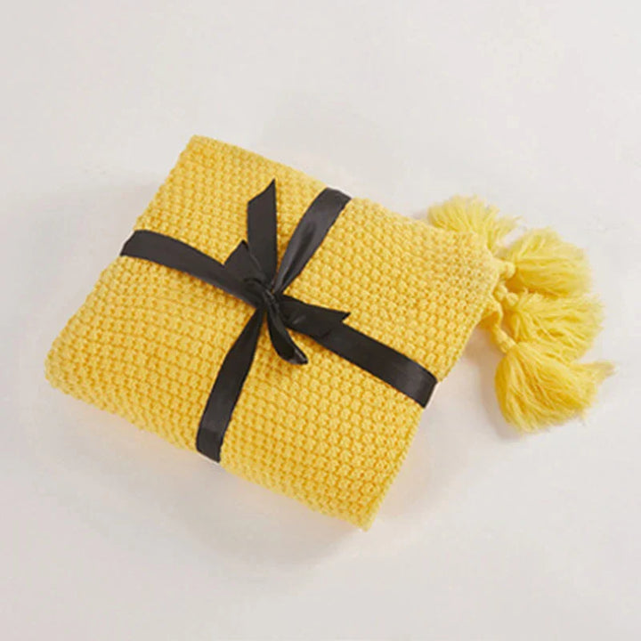 Gelbe Strickdecke mit schwarzen Schleifenband und Quasten, weiche Wohnaccessoires.