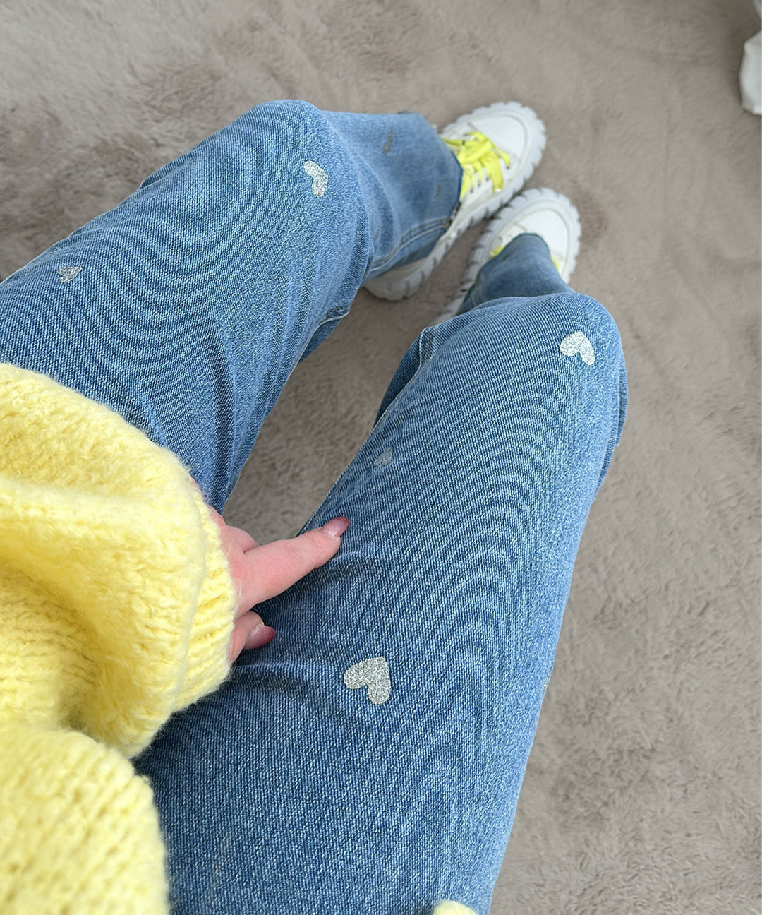 Frau in blauer Jeans mit Herzmuster, gelbem Pullover und weißen Sneakers auf Teppich.