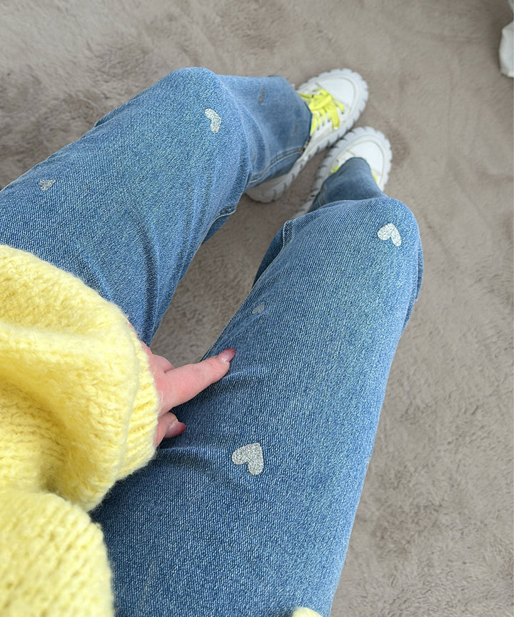 Frau in blauer Jeans mit Herzmuster, gelbem Pullover und weißen Sneakers auf Teppich.