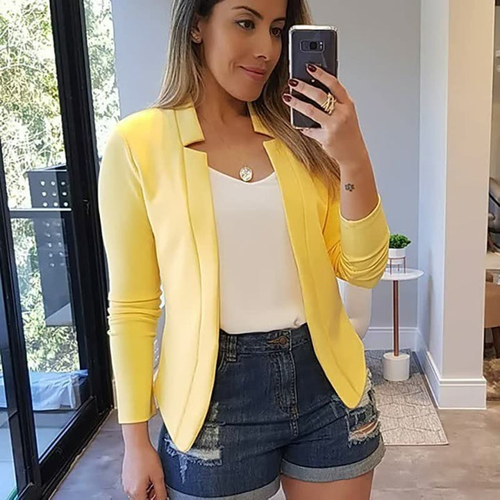 Frau in gelbem Blazer und Jeansshorts macht Selfie im modernen Wohnzimmer.