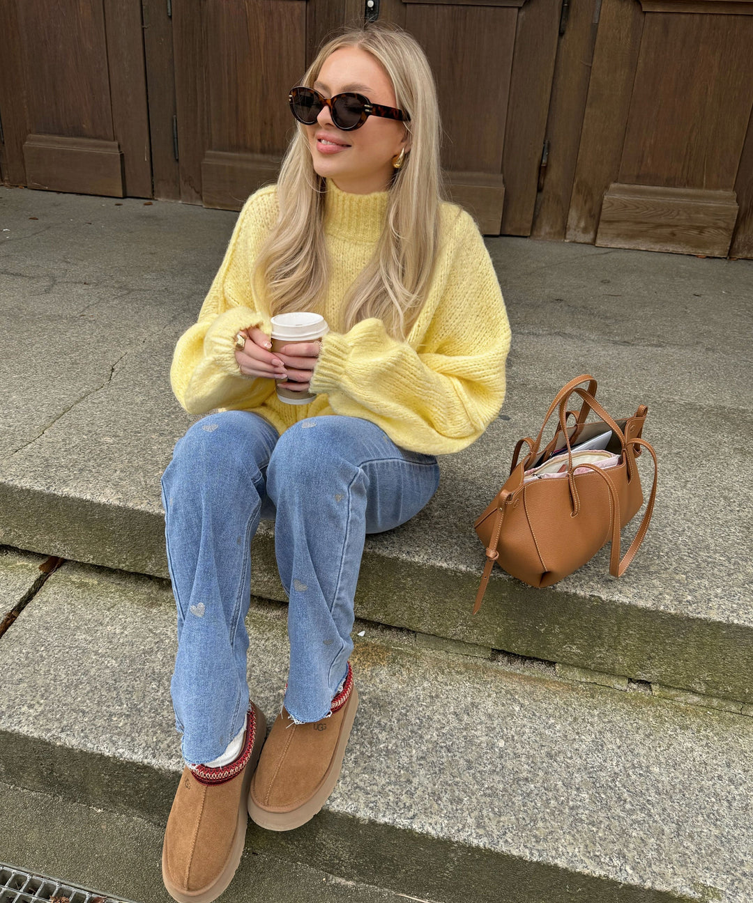 Frau in gelbem Pullover, blauer Jeans, braunen Stiefeln, mit Sonnenbrille und Kaffeetasse.