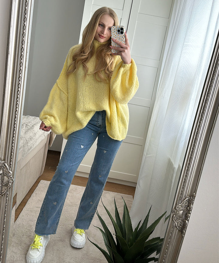 Frau in gelbem Strickpullover und blauer Jeans mit Herzmuster, Selfie im Spiegel.