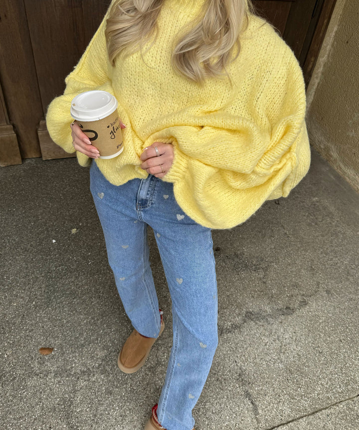 Frau in gelbem Strickpullover, blauer Jeans mit Herzmuster, hält Kaffeebecher, Herbstmode.