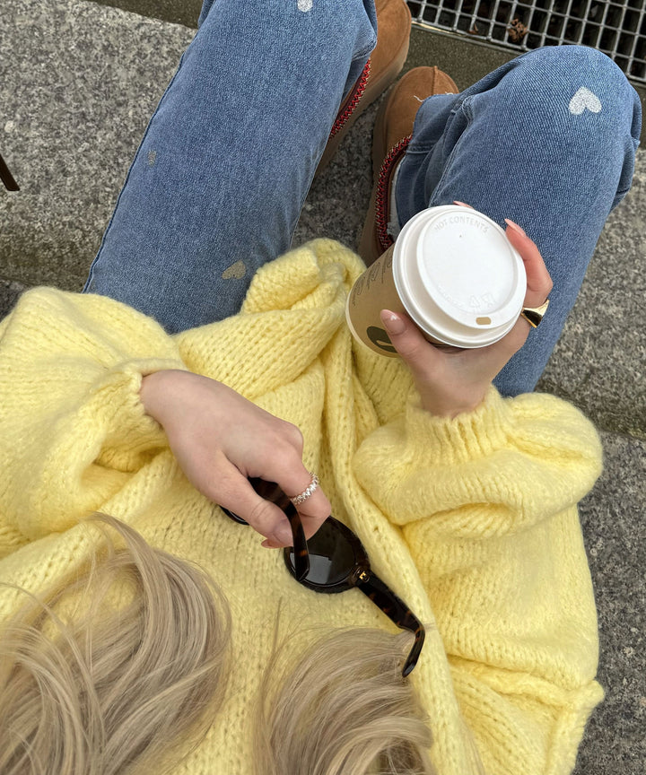 Frau in gelbem Strickpullover hält Kaffeebecher, trägt Jeans mit Herzmuster und Sonnenbrille.
